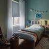 Sheer white grommet curtains flank a lowered grey Roman blind over the window, partially filtering daylight; small teal bedroom with a single bed, bedside lamp, soft toys, and rug.