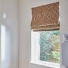 Fabric Roman blind, beige with white diamond pattern, partially lowered over a window, filtering daylight; small stair landing with walls and banister, outside view of shrubs and wooden fence.