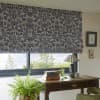 Patterned fabric Roman blind, lowered across the window with dense navy botanical print on cream, filtering daylight over a wooden desk with lamp, books, fern and a treed outside view.