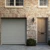 Sheer curtains behind upper windows, drawn to sides and filtering daylight; brick facade with closed roll-up garage door left, wooden front door right, wall lantern and paved driveway.