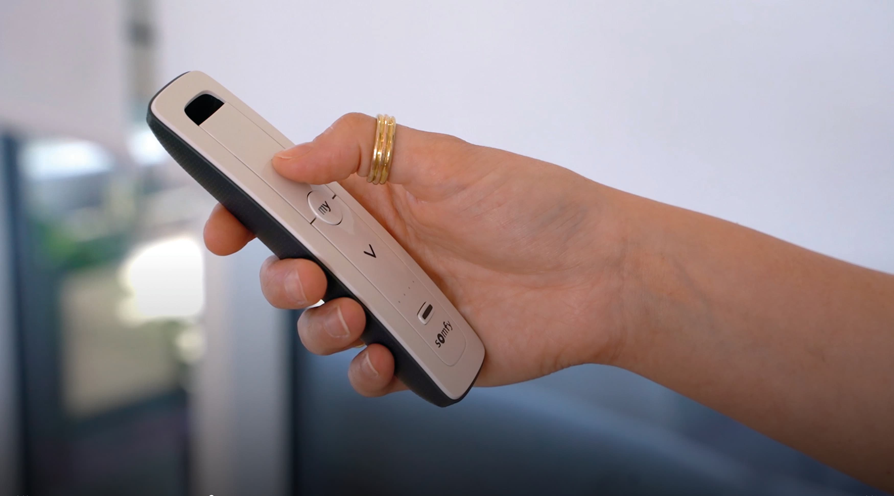 Slim white-and-black remote control held in a right hand, thumb pressing a circular my button; indoor, softly lit room with a blurred window or glass door in the background.
Text: my V somfy