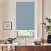 A blue patterned roller blind covers a window above a kitchen sink, partially lowered; the countertop holds a bowl of leafy greens, potted plants, a faucet, and a framed sketch on the wall.