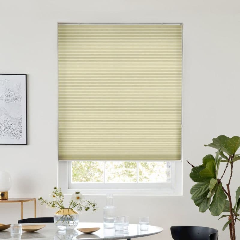 Pale-yellow cellular shade, lowered over a window with a small gap at the bottom, filtering daylight; modern dining area with white walls, round table, glassware and a potted plant.