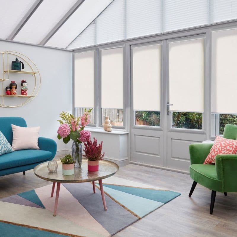 Cream roller blinds covering upper portions of multiple tall glass panes and door, lowered to mid-height to filter light; bright conservatory with teal sofa, green armchair, coffee table and flowers.