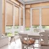 Honey colored horizontal Venetian blinds on tall bay windows, lowered and filtering daylight; white dining table and chairs in a sunlit breakfast room, small stool and green garden beyond.