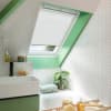 White pleated skylight blind, mostly lowered over a roof window, filtering daylight; bathroom with green-painted sloped ceiling, white herringbone tiles, a sink and tub, plant, and toiletries.