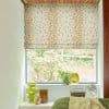 Patterned Roman blind (beige with scattered brown dots) lowered halfway, filtering daylight; bedroom window sill with green vases, potted plant, fan and radio, bed with green knit blanket, garden view.