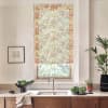A floral roller blind filters sunlight over a kitchen sink; wooden countertop holds a bowl of greens, potted plants, framed sketch on the wall, and a tall leafy plant to the right.
