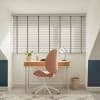 Horizontal slatted blinds over a wide window, pale grey with tapes and cord, closed and filtering daylight, casting striped shadows across a study with wooden desk and pink swivel chair.