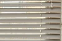 Horizontal Venetian blinds with wide grey slats and a vertical bead chain, partially closed, filtering soft daylight into an interior, narrow bright gaps revealing an exterior light.