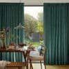 Thick teal woven floor-length curtains, parted centrally to reveal a sunlit glass door; dining table with breakfast plates, pendant lamp, wooden chairs, opening onto a green garden.