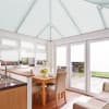 Pleated white conservatory blinds—roof panels and window/door coverings—closed and filtering soft daylight; in a bright dining conservatory with wooden table, beige chairs, kitchen sink, and patio garden beyond.