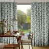 Pleated teal-gray geometric curtains, full-length, frame a central open sliding door, parted to admit sunlight; a dining table set for breakfast overlooks a garden with trees, swing, and shed.