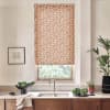 Patterned floral roller blind covers a window above a stainless-steel sink. Sunlight filters through; a wooden countertop holds a bowl of produce, potted plants, a towel, and a framed sketch.