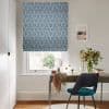 Blue leaf-patterned Roman blind, lowered and filtering daylight; fitted over a sash window. Context: bright home office with wooden desk, teal upholstered chair, vase of flowers, mirror and small decor.