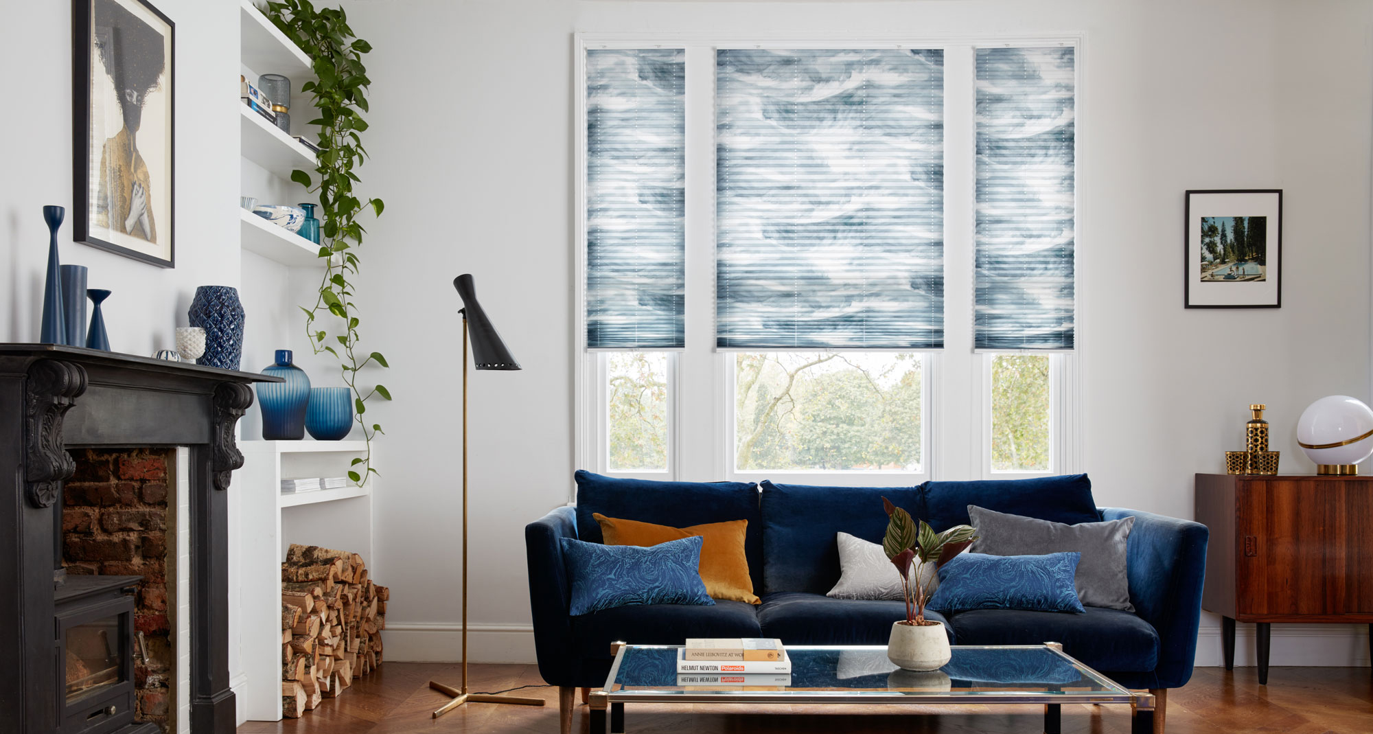 Blue and white shaded Pleated blinds dressed on windows of living room. There is blue sofa decorated with blue, grey and mustard cushions have been placed in the room.